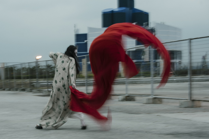 Eiko in a kimono with her back towards the camera, her red scarf flaps violently in the wind